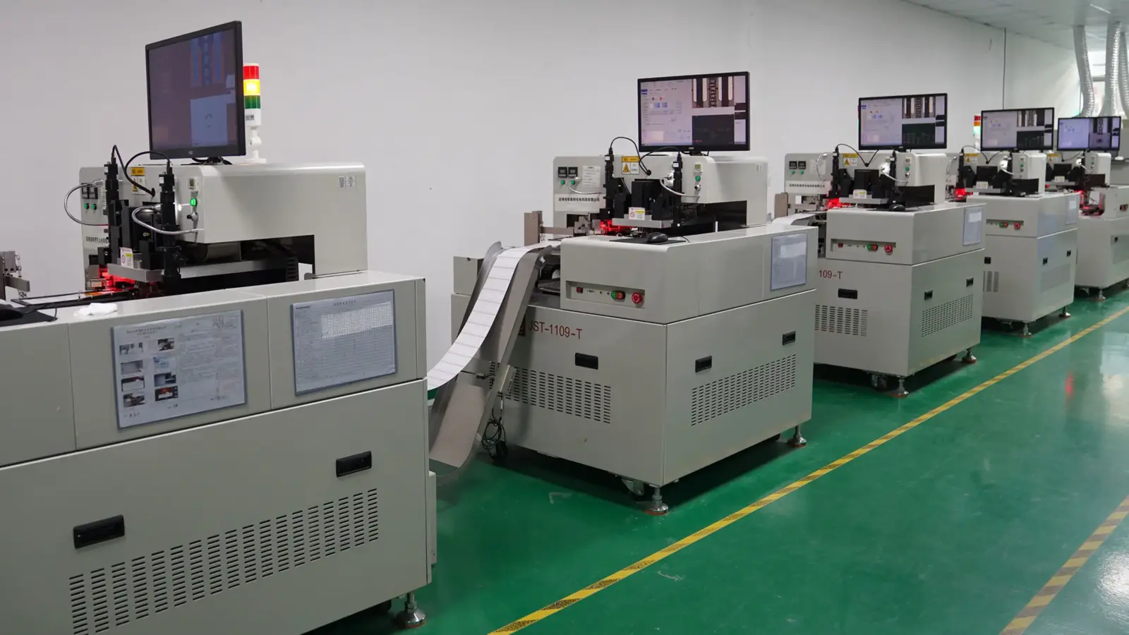 Row of automated LED strip testing machines with monitors and signal towers, flexible strip feeding through each unit