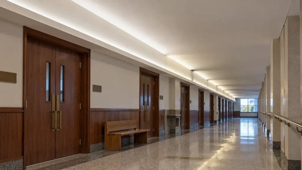 County courthouse public corridor cove with the left half still lit by a T5 fixture row showing dark socket gaps and the right half lit by one continuous LED channel at 4000K