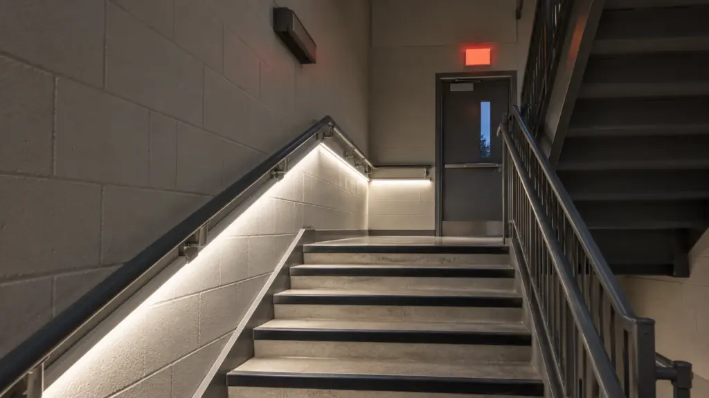 High school exit stair landing lit by 120V LED strip on the emergency circuit, the strip hidden under the handrail line and the wall fixtures off